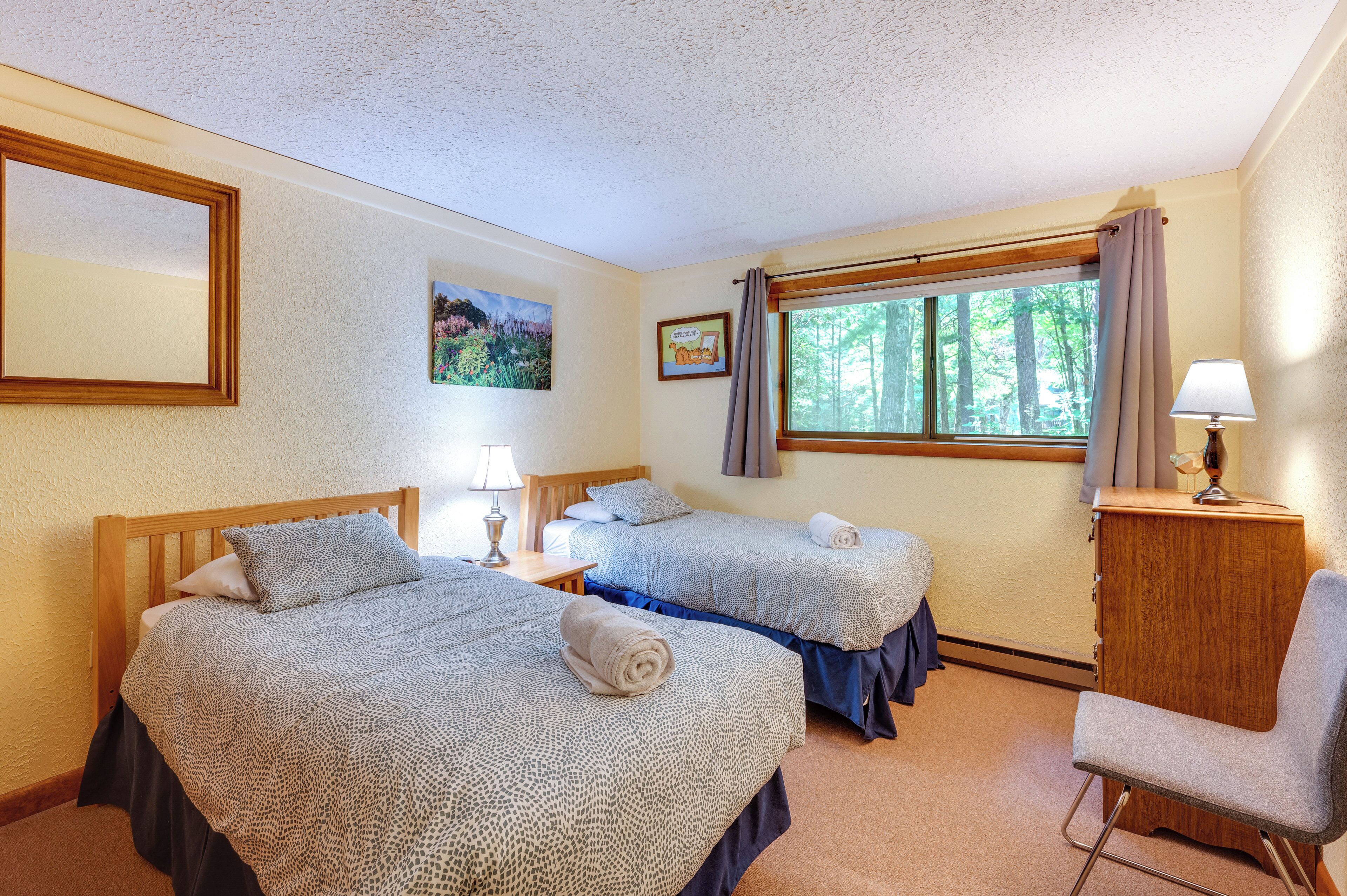A hotel room with two beds, a desk, a chair, a window with curtains, and a lamp.