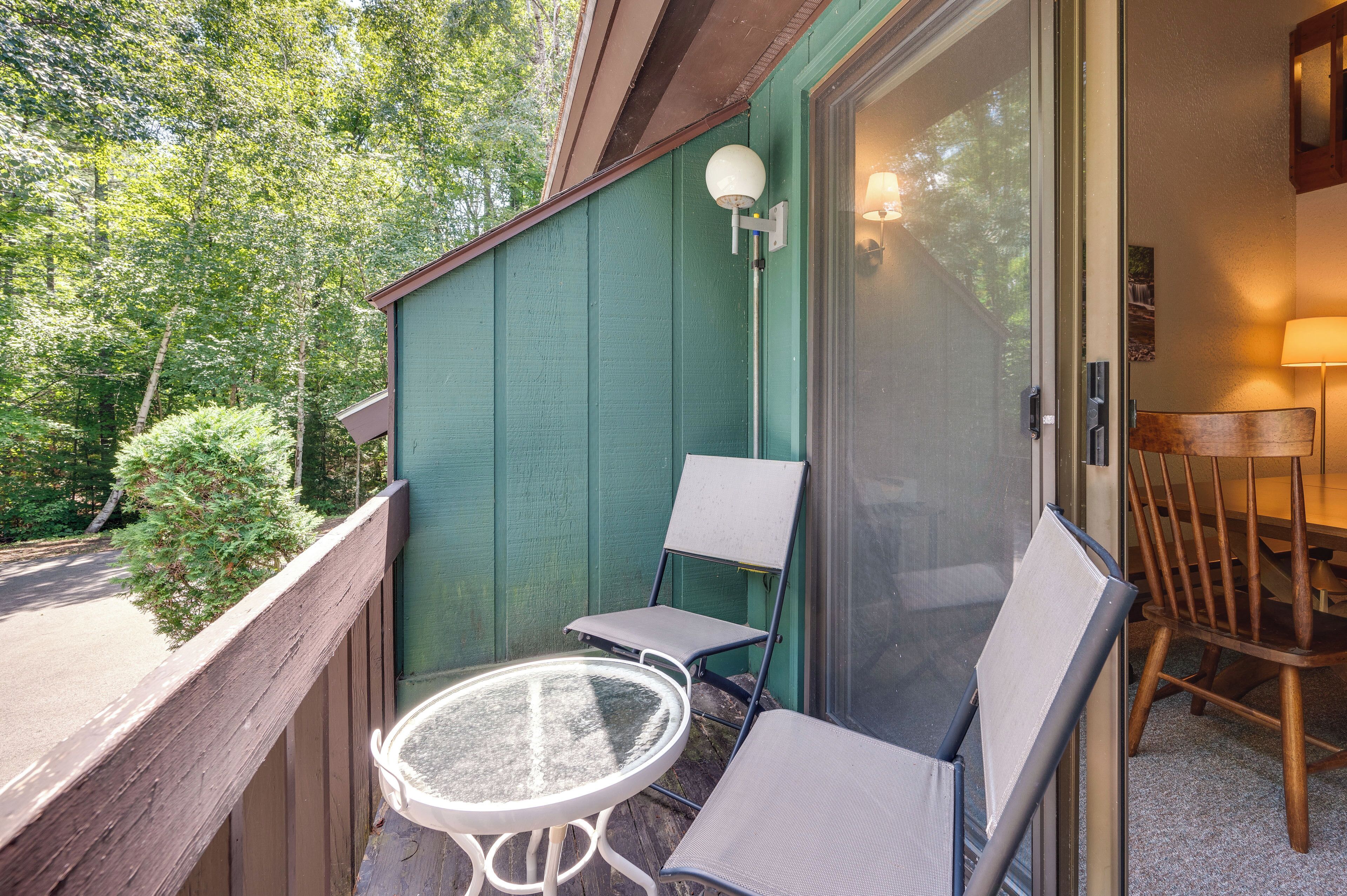 A balcony with a glass table and two chairs, a wooden chair, and a lamp.