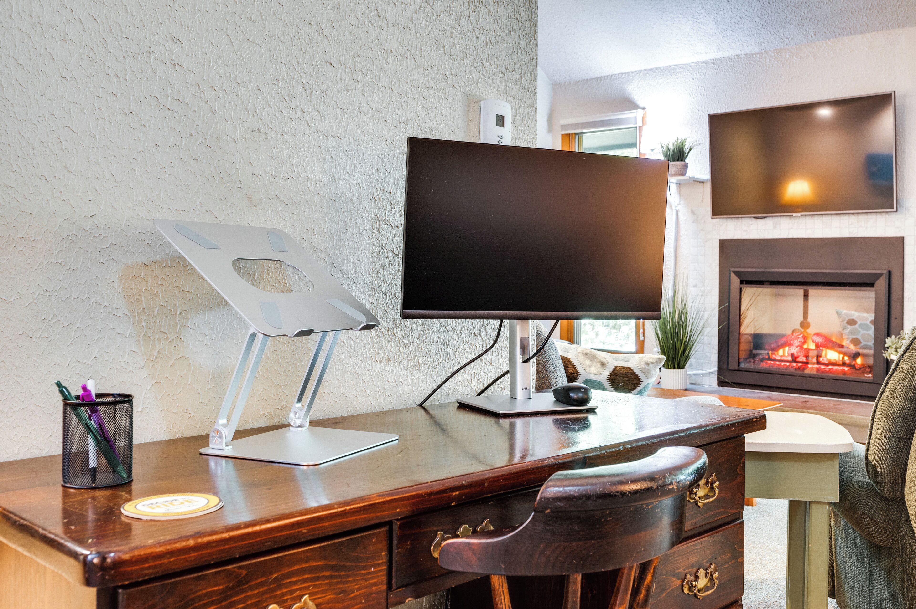 A modern office space with a wooden desk, a computer monitor, a fireplace, and a television mounted on the wall.