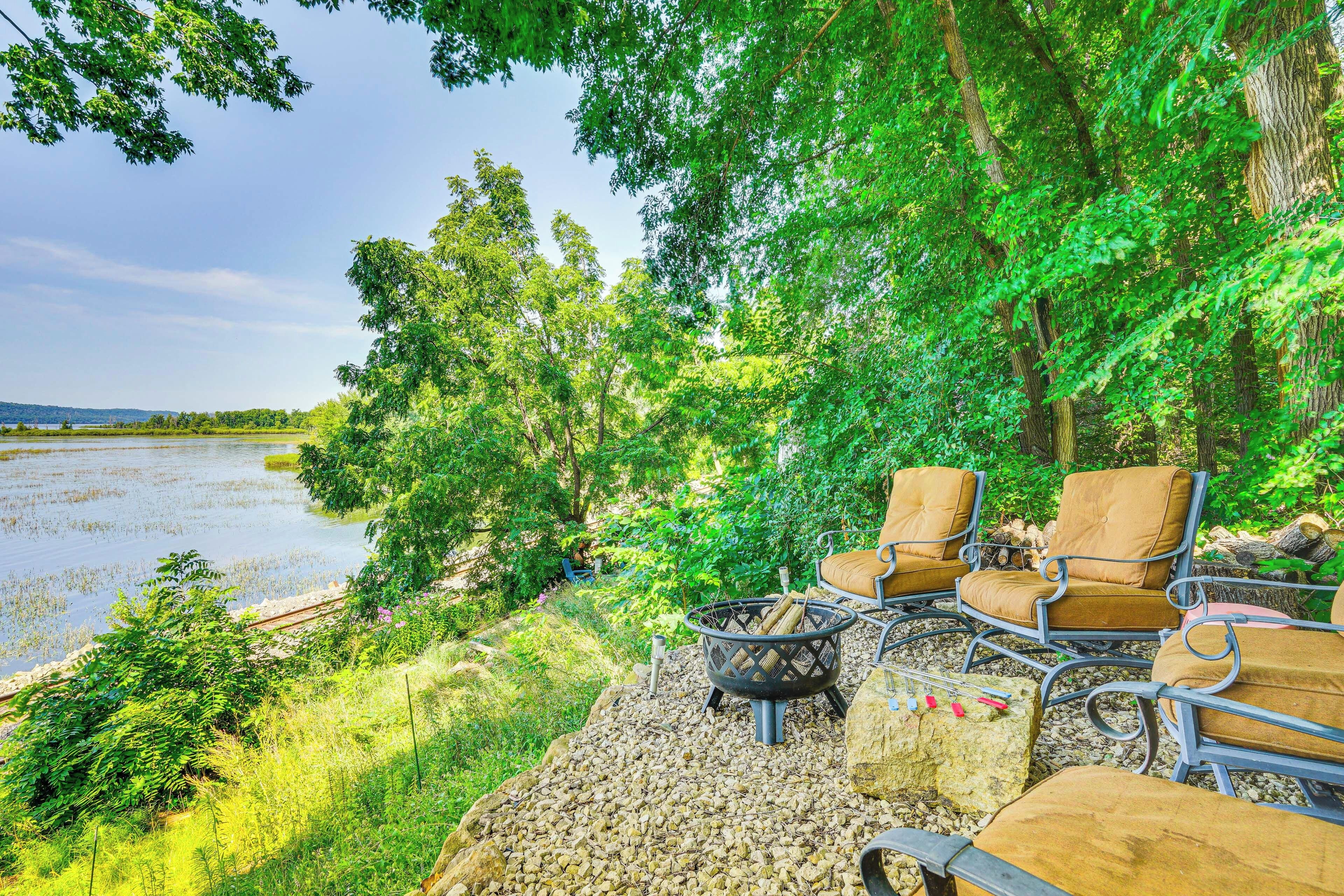 A lakeside seating area with chairs and a fire pit, surrounded by lush greenery and a view of the lake.