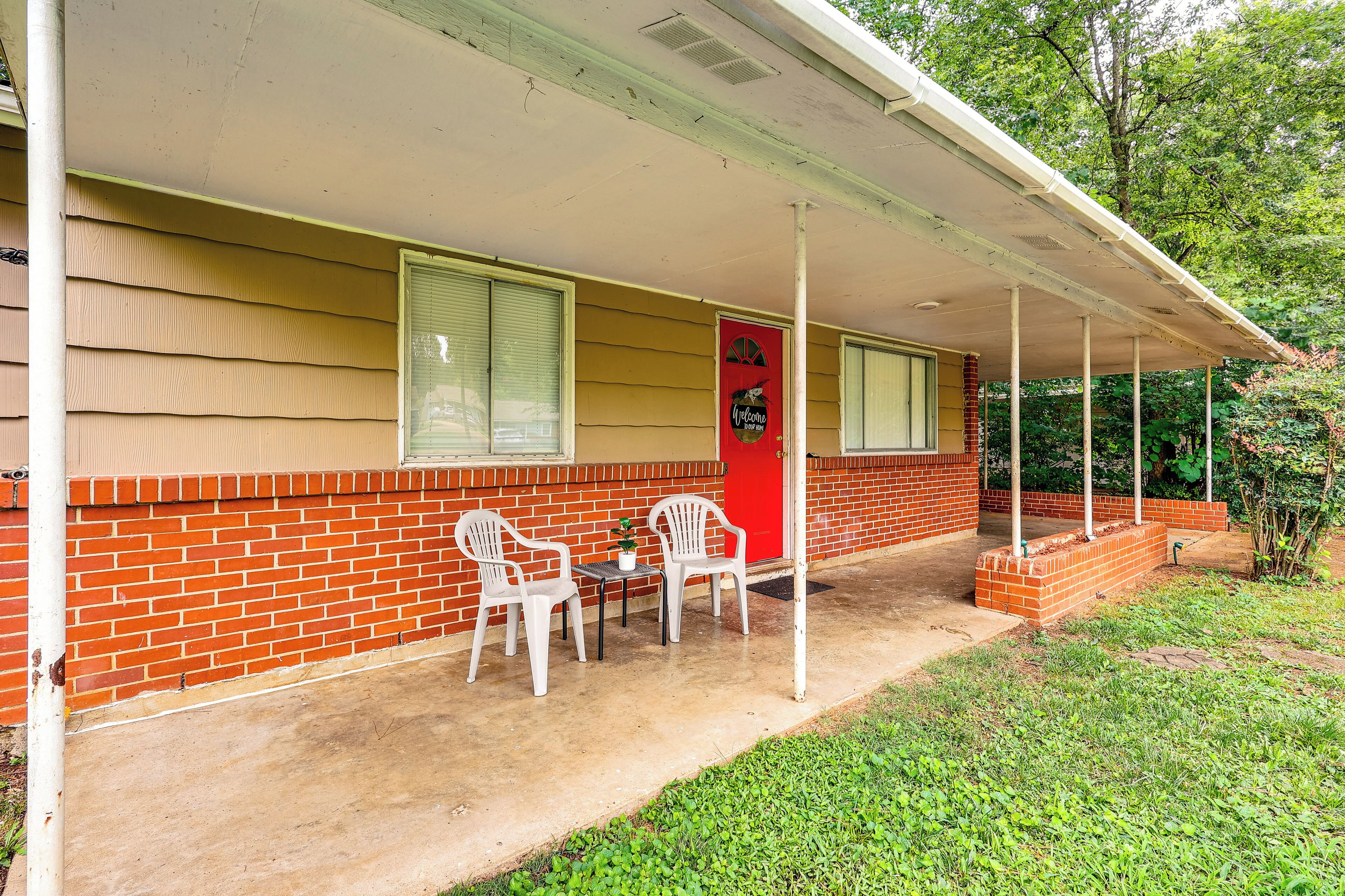 A red door with a welcome sign, a covered porch with two white plastic chairs, and a brick wall.