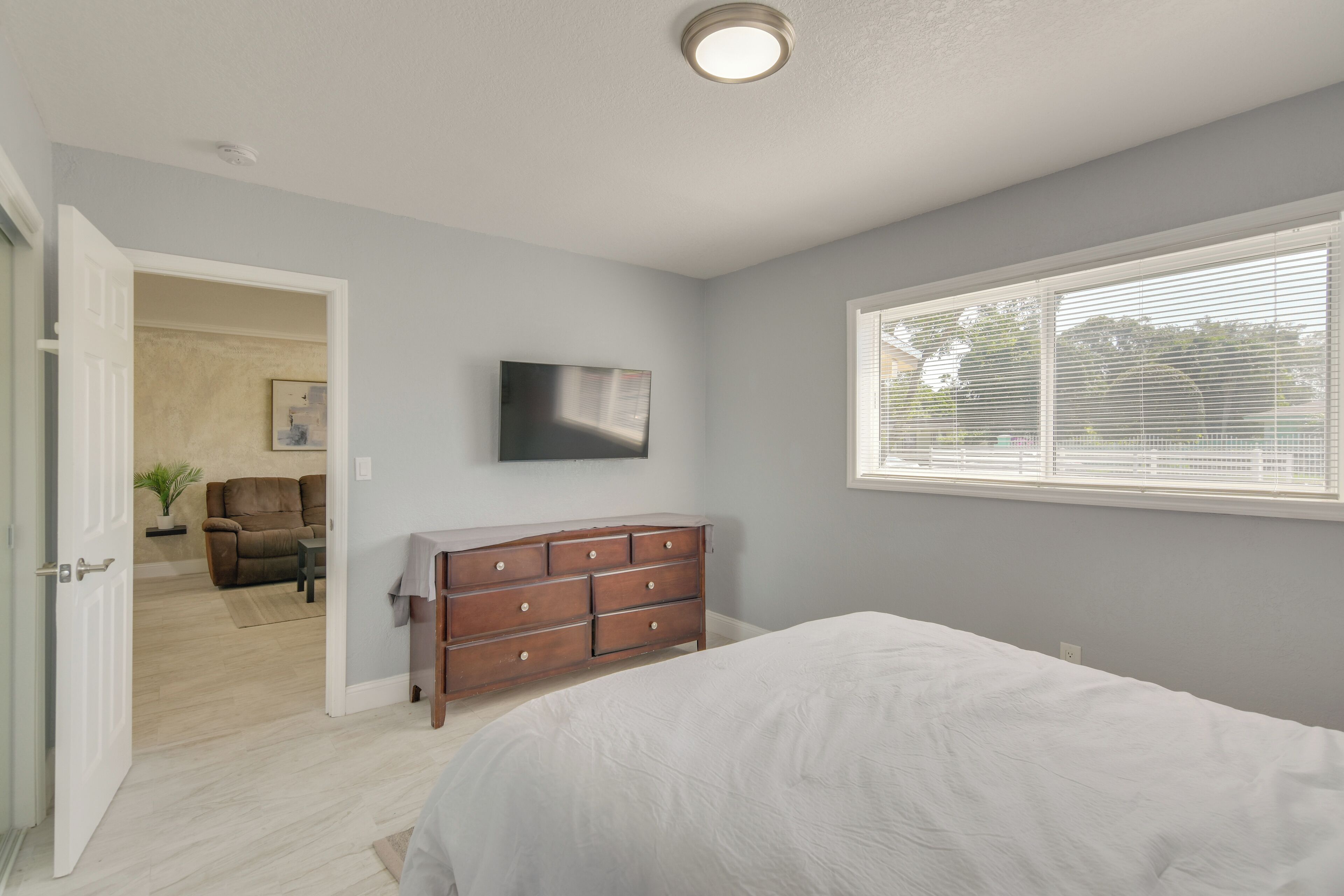 Une chambre à coucher avec un lit, une commode, un téléviseur fixé au mur et une fenêtre avec des stores.