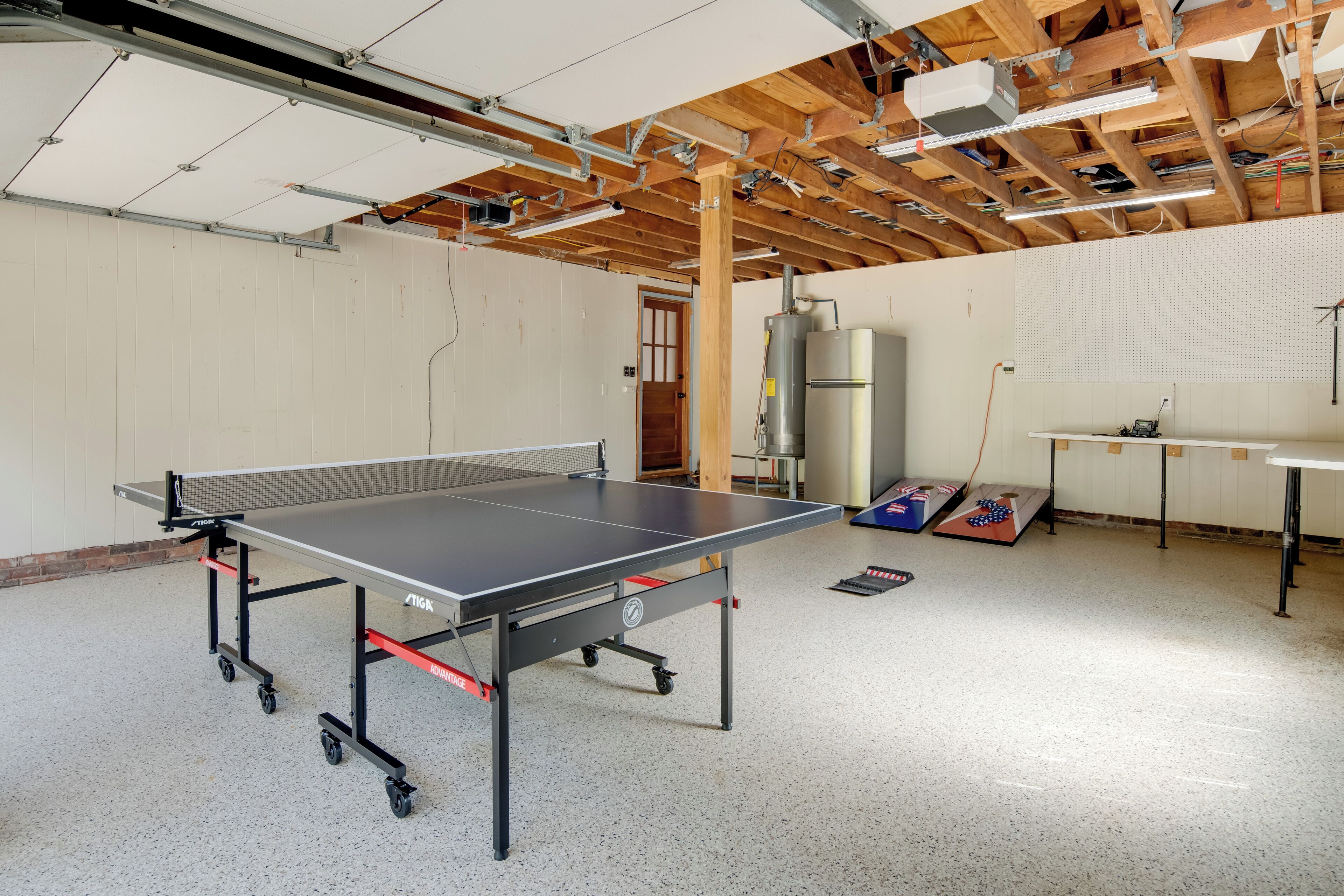 Una mesa de ping pong en una habitación con vigas de madera a la vista, un panel perforado y un calentador de agua grande.