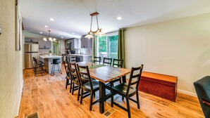 A spacious dining area with a wooden table and chairs, a kitchen in the background, and a view of greenery outside.