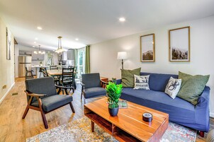 A modern living room with a blue sofa, wooden coffee table, and chairs. There is a dining area with a table and chairs, and a kitchen in the background.