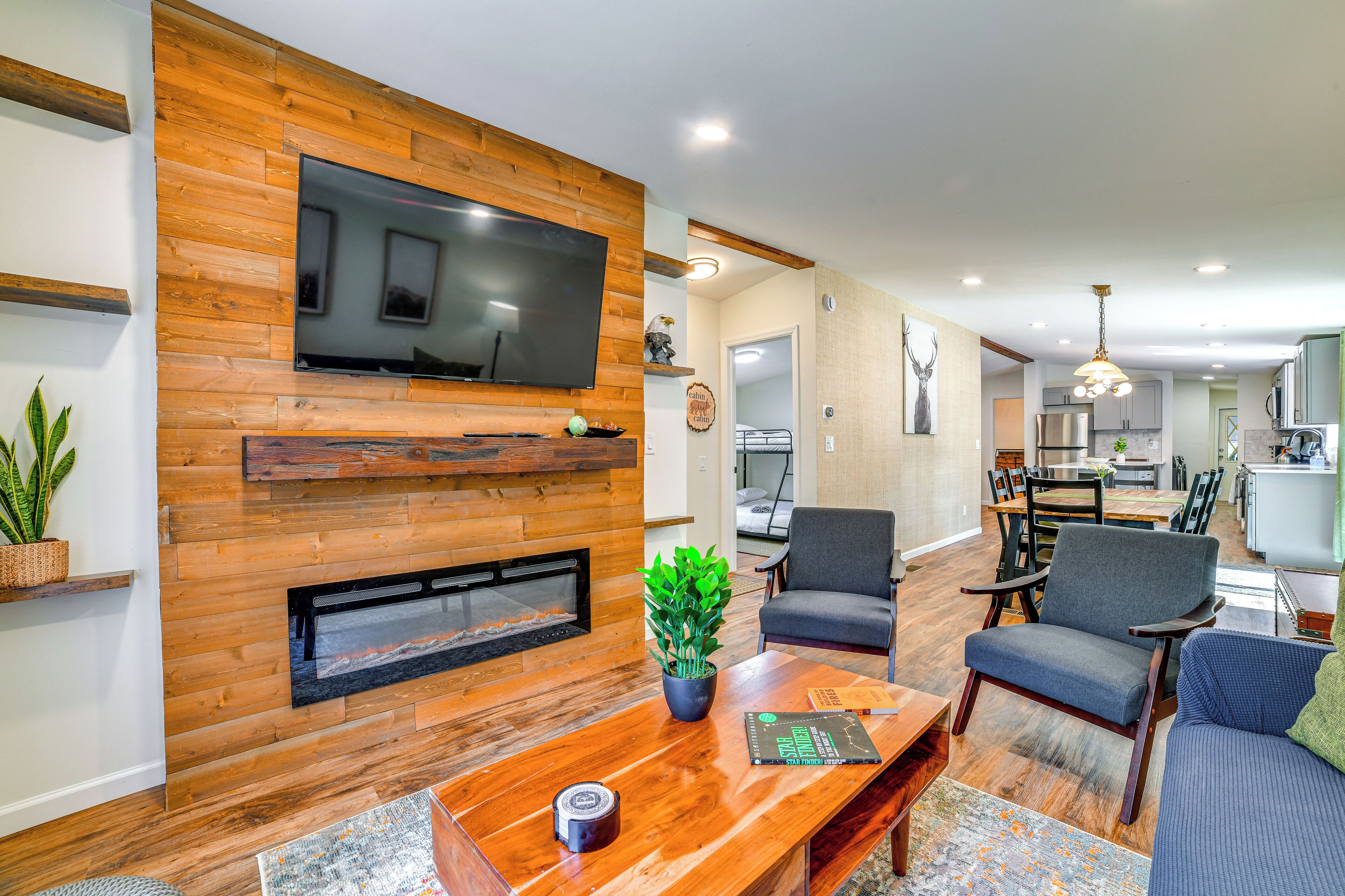 A modern living room with a fireplace, a flat-screen TV, and a dining area with a kitchen in the background.