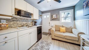 A modern kitchen with white cabinets, a black microwave, and a wooden backsplash. There is a beige sofa with a yellow pillow and a small table with a lamp. - Charming Geneva Vacation Rental ~ 5 Mi to Lake! (Geneva)