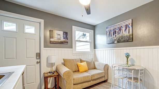 A cozy living area with a beige sofa, a small side table, a white table with two chairs, a window, and a wall-mounted artwork. - Charming Geneva Vacation Rental ~ 5 Mi to Lake! (Geneva)