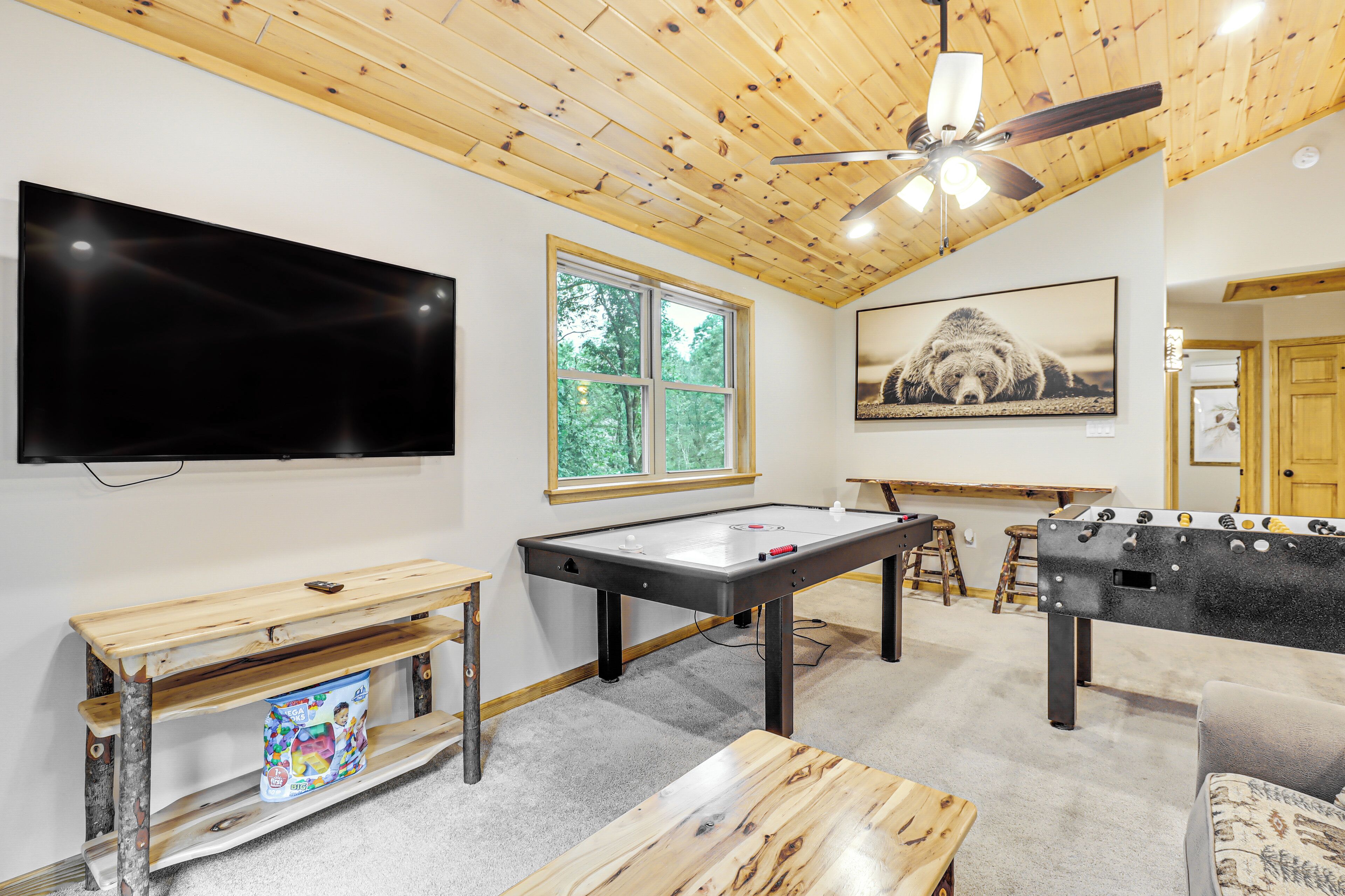 A room with a foosball table, a flat-screen TV, a ceiling fan, and a large window.