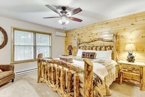 Une chambre à coucher avec une tête de lit en bois, un lit recouvert d’une couette blanche, une table de chevet avec une lampe et une fenêtre avec des stores.