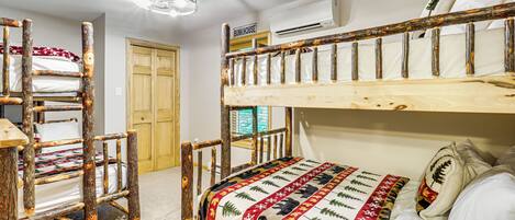 Una cama litera con estructura de madera y colcha acolchada. Se observa una silla de madera con cojín, una puerta de madera y un equipo de aire acondicionado en la pared.