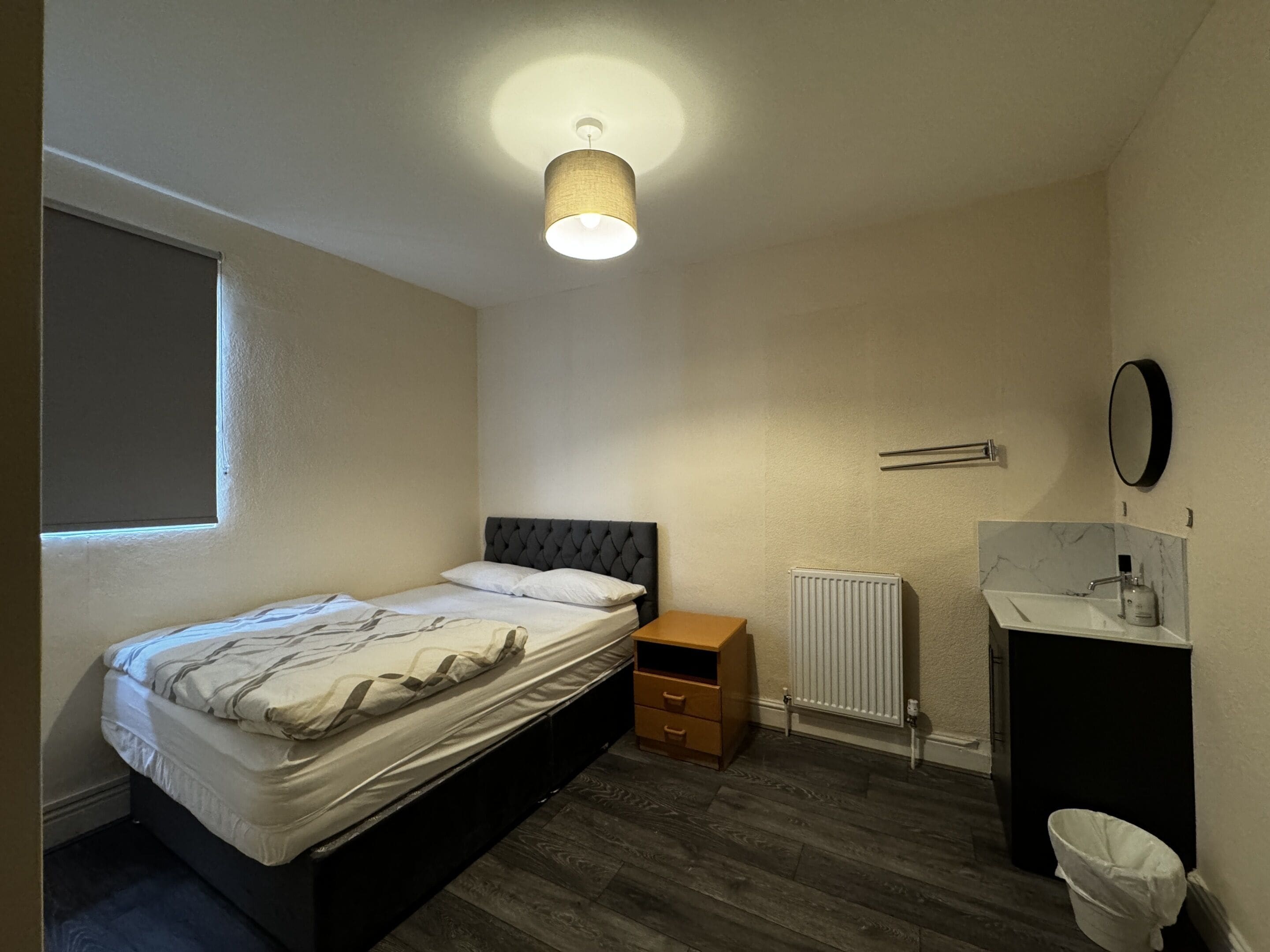 A bedroom with a bed, a bedside table, a radiator, a mirror, and a window.