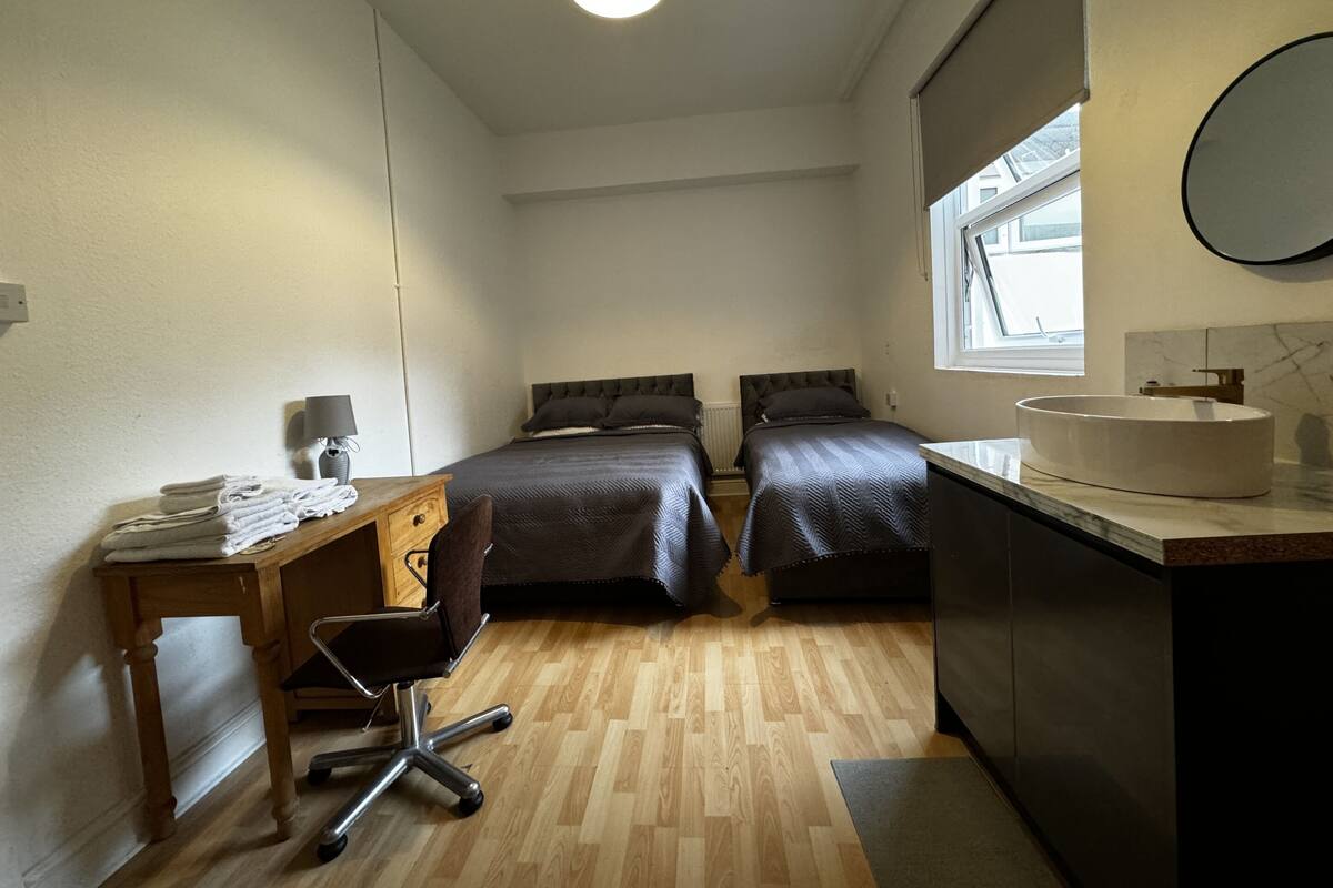 Una habitación pequeña con dos camas, un escritorio con silla, un lavamanos y una ventana.
