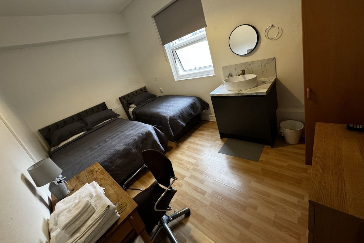 Una habitación compacta con dos camas, un escritorio, una silla, un baño pequeño con lavamanos y una ventana que deja entrar luz natural.