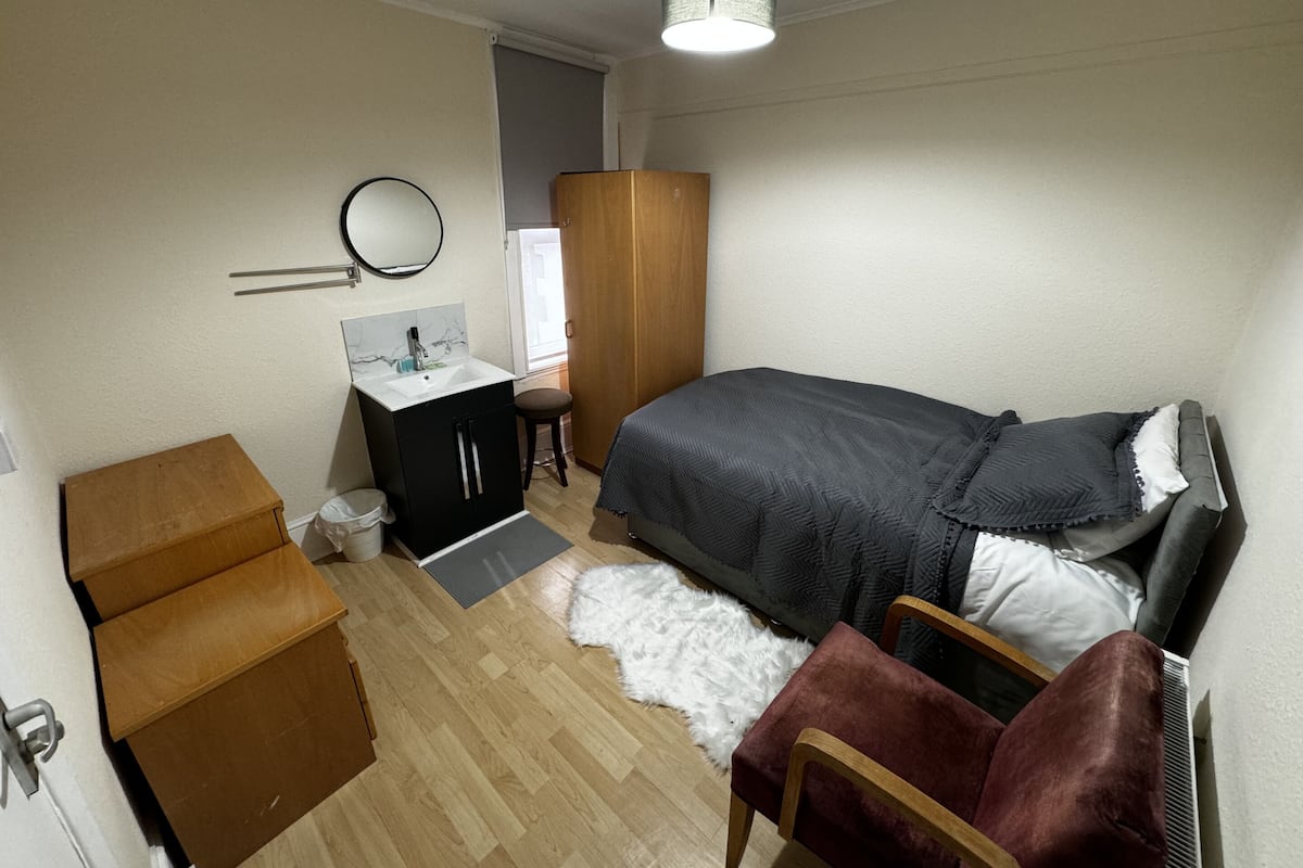 Un dormitorio con cama, un armario de madera, un escritorio pequeño con silla, un espejo y una lámpara.