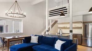 Ein modernes Wohnzimmer mit einem blauen Ecksofa, einem Holzessttisch und einer Chandelier. Im Hintergrund ist eine Küche mit einem Edelstahlkühlschrank und einer Treppe zu sehen.