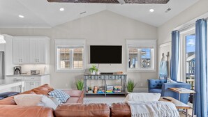 A spacious living room with a brown leather sofa, a flat-screen TV mounted on the wall, and a dining area with white cabinets and a kitchen in the background. - LinkedOUT #171 – 3 BD Dog Friendly Greensboro St Duplex Steps from the beach! (Wrightsville Beach)