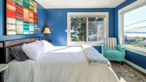 A bedroom with a large bed, a window with a view, a colorful abstract painting, and a teal chair. - Orca Haven: 2BR w/ Scenic Views, Richmond Beach (Shoreline)