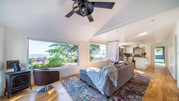 A spacious living room with a large window, a fireplace, a sofa, and a ceiling fan. - Orca Haven: 2BR w/ Scenic Views, Richmond Beach (Shoreline)