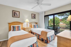 A hotel room with two beds, a ceiling fan, a window with a view of greenery, and a table with chairs outside.