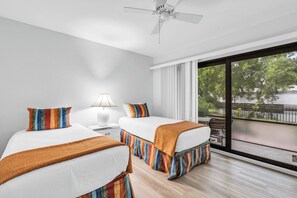 A hotel room with two beds, a ceiling fan, a nightstand with a lamp, and a large window with a view of trees and a balcony.