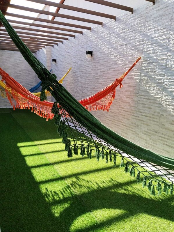 A hammock suspended between two wooden beams in a room with a white brick wall and artificial grass flooring. - Pousada Nelly Medeiros (Carolina)
