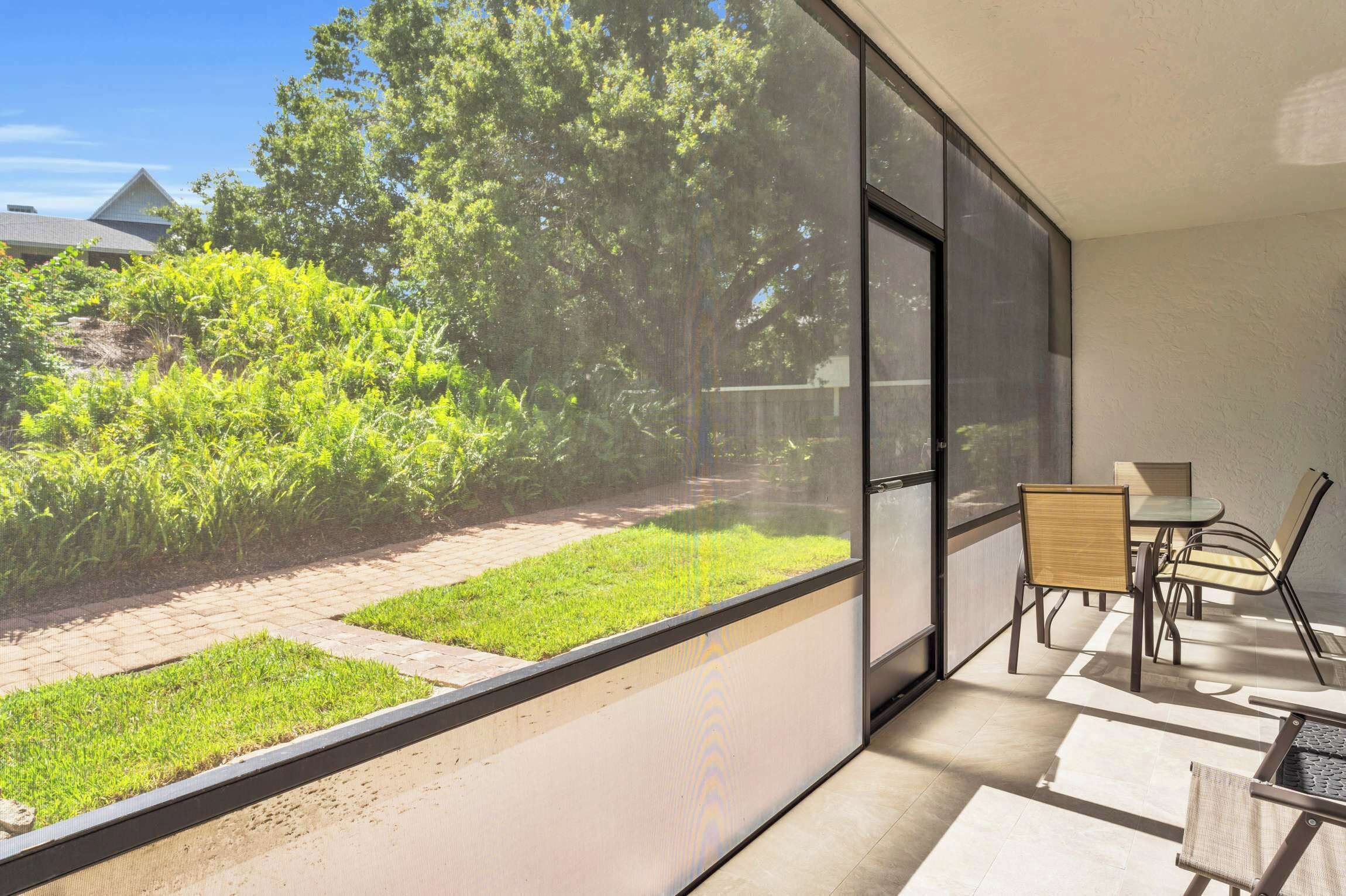 A modern balcony with a glass wall, a small table, two chairs, and a view of a garden and trees.