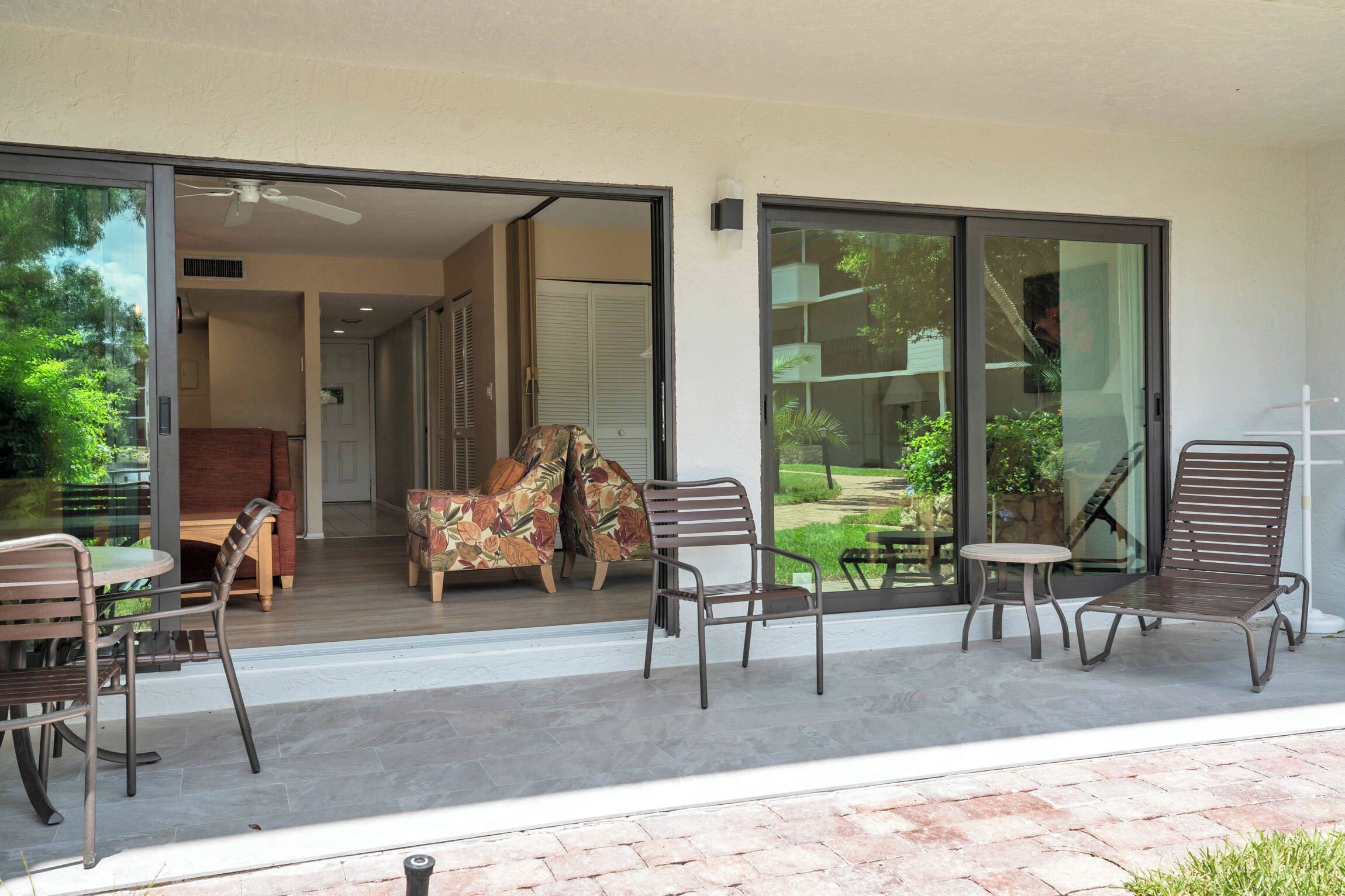 A modern outdoor patio with a glass door leading to a living area, furnished with a sofa, chairs, and a small table.