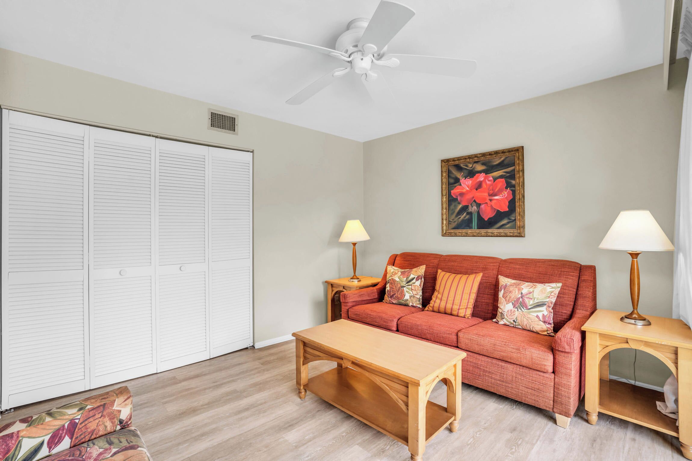 A living room with a red sofa, two side tables with lamps, a wooden coffee table, a ceiling fan, and a large floral painting on the wall.