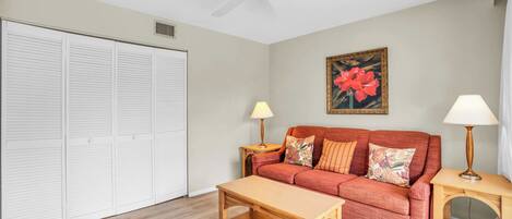 A living room with a red sofa, two side tables with lamps, a wooden coffee table, a ceiling fan, and a large floral painting on the wall.