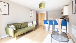 Ein modernes Wohnzimmer mit einem grünen Sofa, einem Glastisch und einer Küchenzeile mit blauen Barhocker.