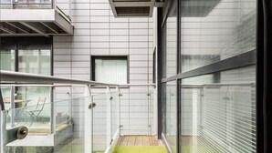 Ein modernes Gebäude mit einem Glasbalkon, einer kleinen, abgegrenzten Fläche mit künstlichem Rasen und einem Fenster mit Jalousien.