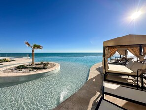 A clear pool with a view of the sea, a palm tree, and a covered lounge area with chairs. - 3BR Ground Floor Condo With Direct Pool Access (Puerto Peñasco)