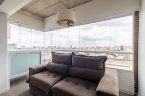 A modern living room with a large window offering a city view, a brown sofa, and a hanging light fixture.