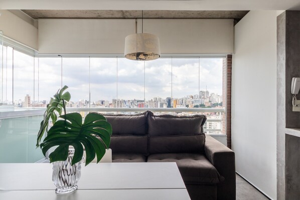 A modern living room with a large window, a brown leather sofa, a white table, and a clear vase with a plant.