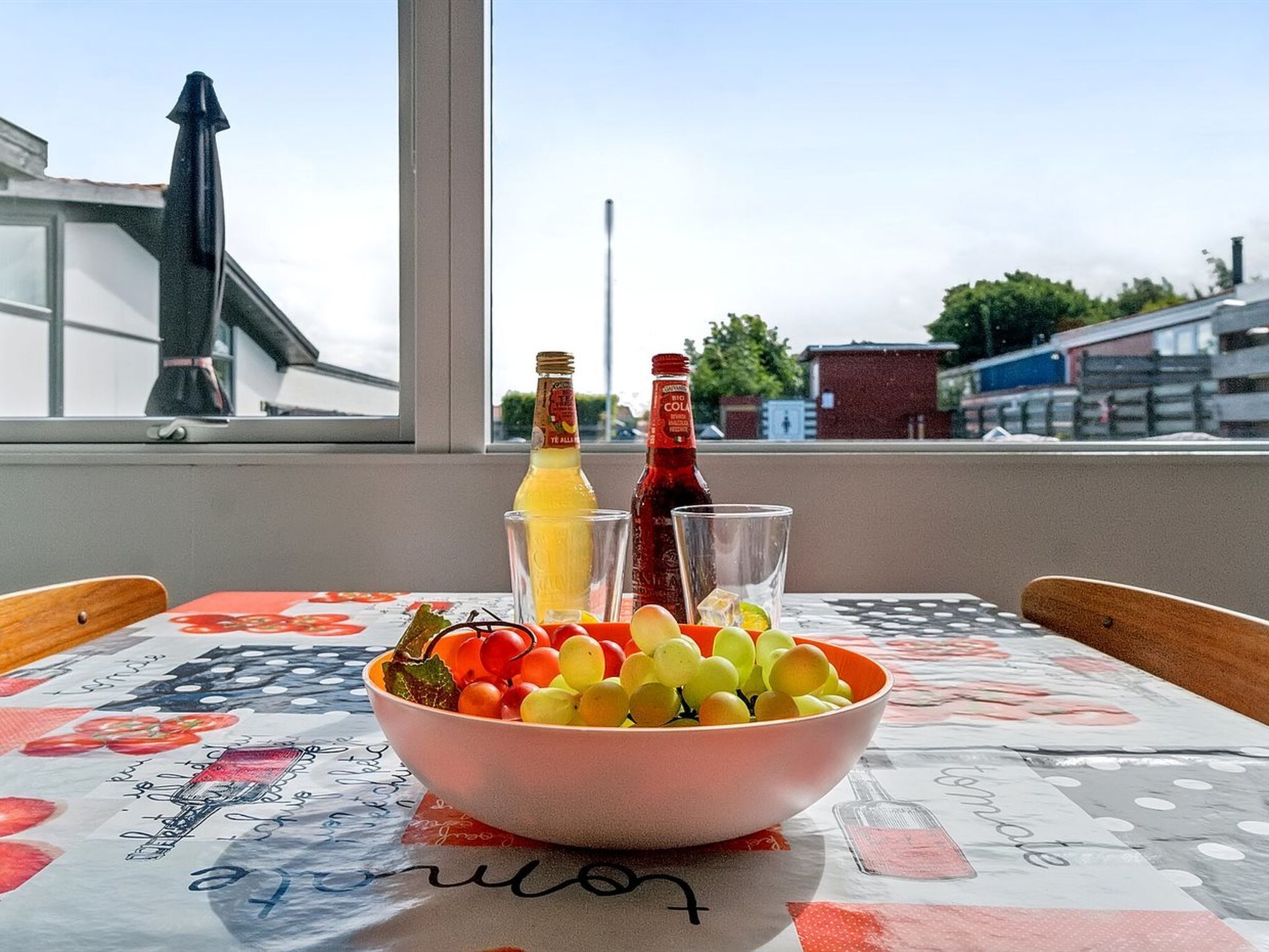 A table with a bowl of fruit, two bottles, and glasses on a tablecloth with a view of buildings and a chimney in the background.