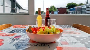 Una mesa con un bol de fruta, dos botellas y vasos sobre un mantel, con vistas a edificios y una chimenea al fondo.