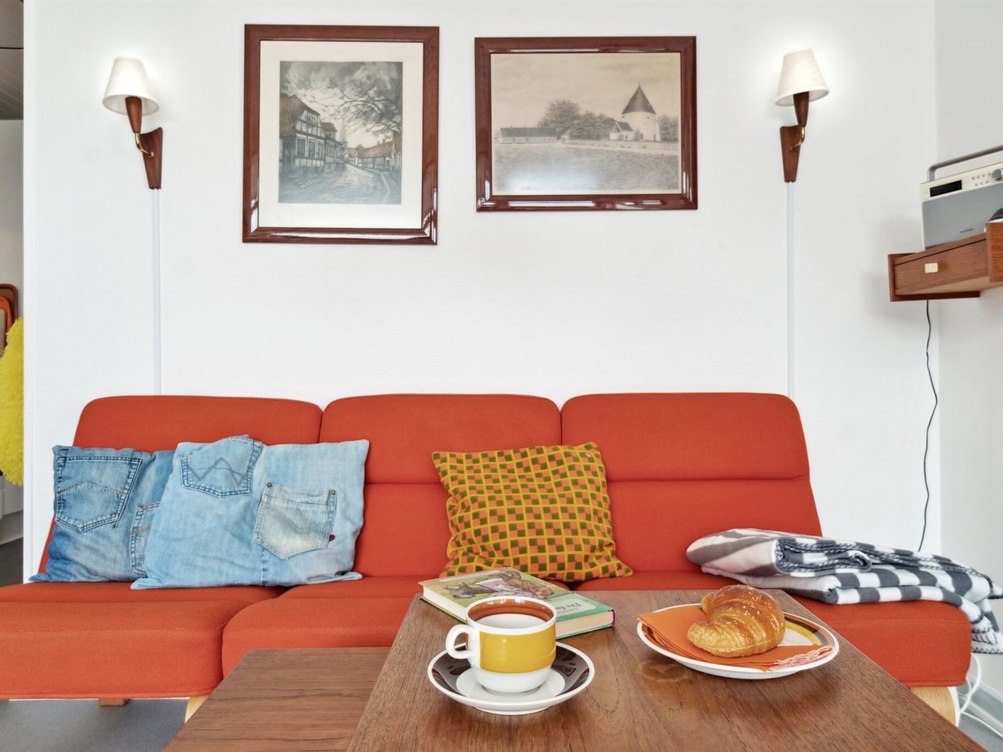 Een gezellige woonkamer met een rode bank, twee ingelijste foto’s, een bijzettafel met een kopje en croissants, en een lamp aan de muur.
