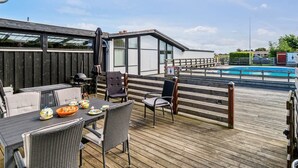 Una terraza de madera con mobiliario exterior, piscina y un edificio con amplios ventanales.
