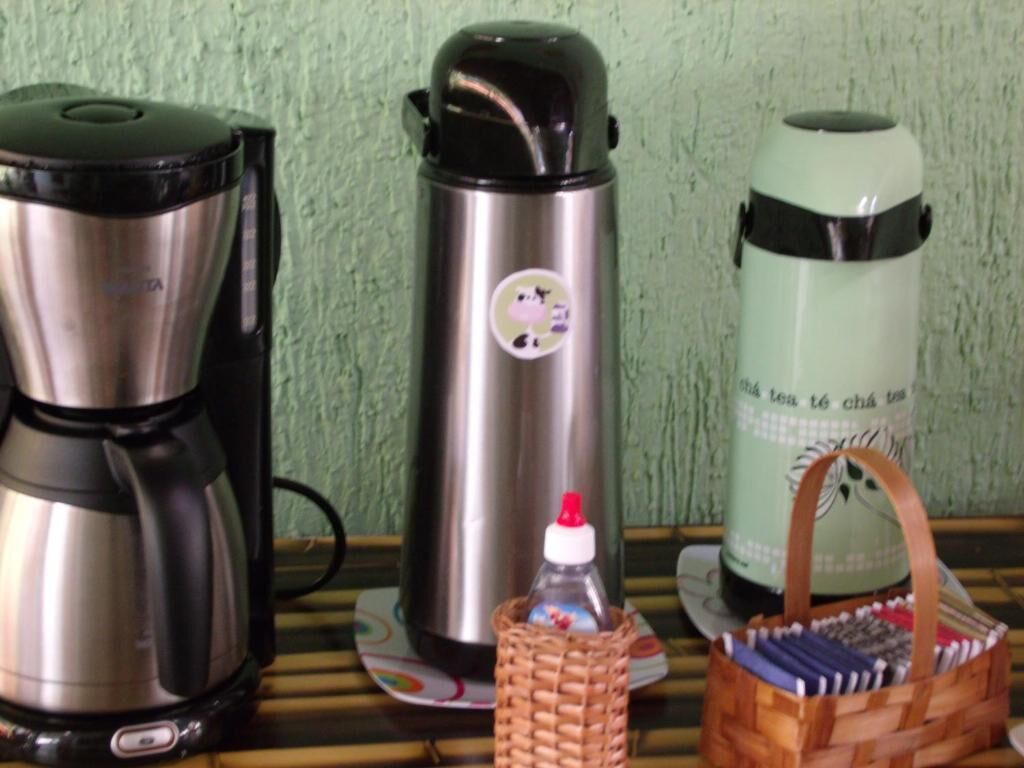 Uma cafeteira de aço inoxidável, uma garrafa térmica com um adesivo de vaca e um recipiente isolante verde sobre um tapete de bambu.