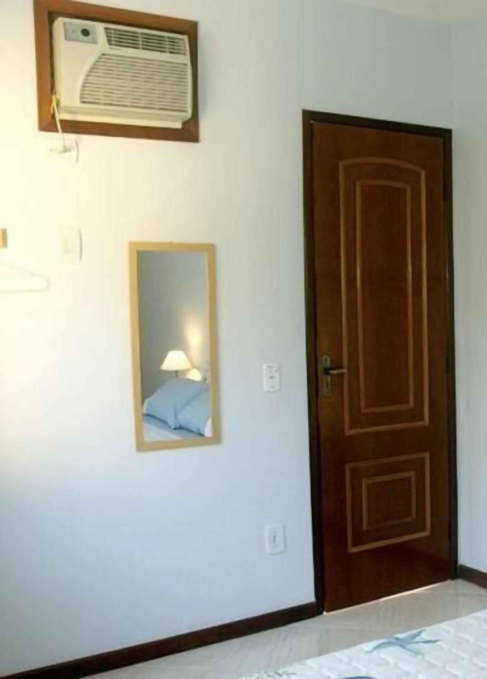 A room with a wooden door, a wall-mounted air conditioner, and a framed mirror reflecting a bed with a lamp.