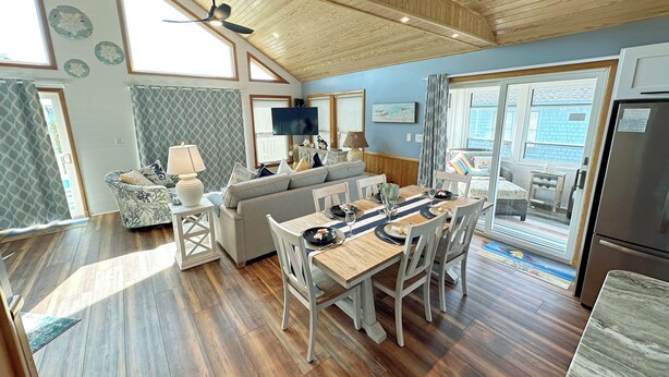 A spacious living area with a high ceiling, wooden flooring, a dining table with chairs, a sofa, and a television. - Oceanside home in Corolla with 4 bedrooms, 3 baths and private pool. (Corolla)