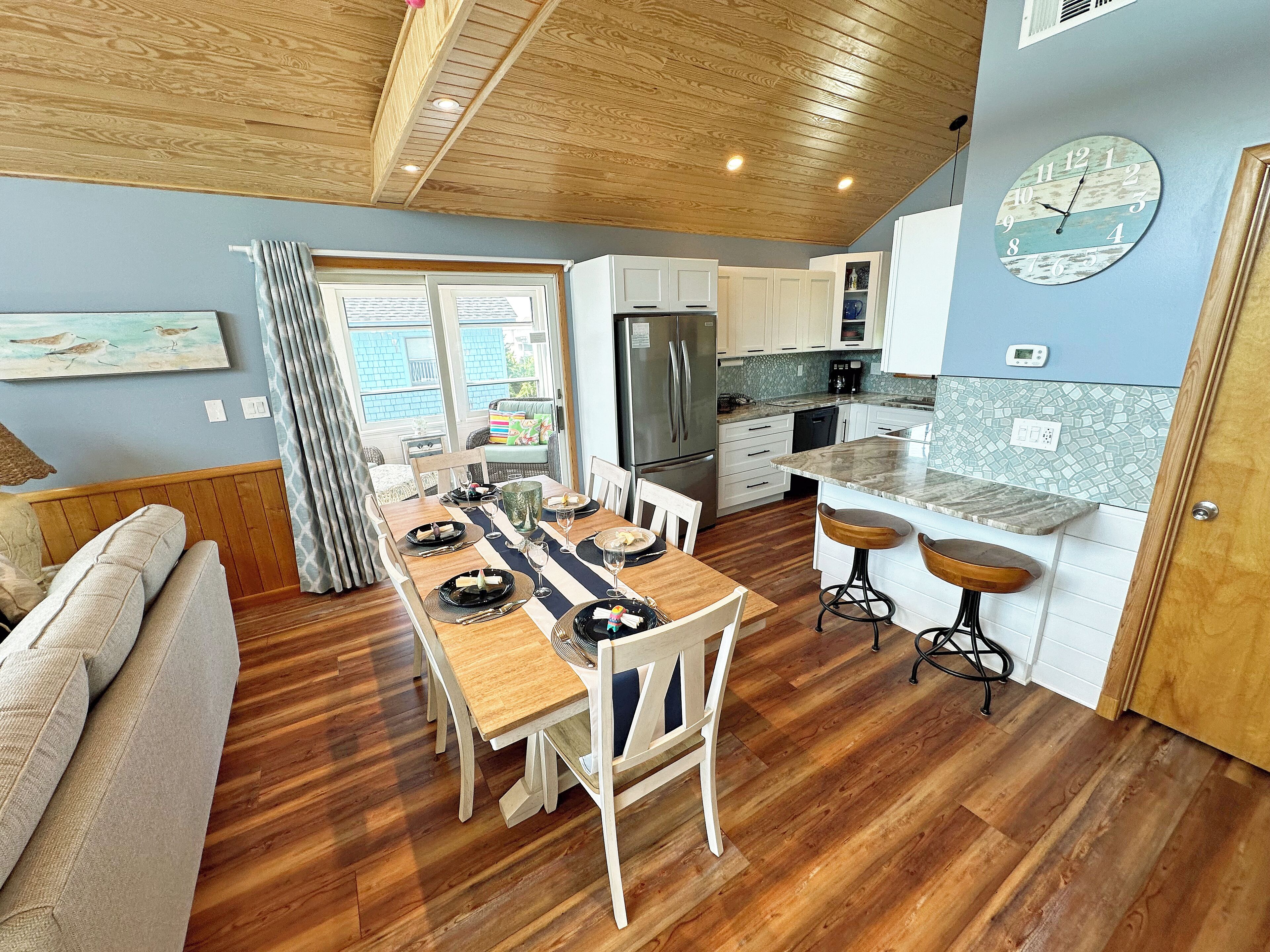 A modern kitchen with white cabinets, a wooden dining table set for four, and a large clock on the wall.