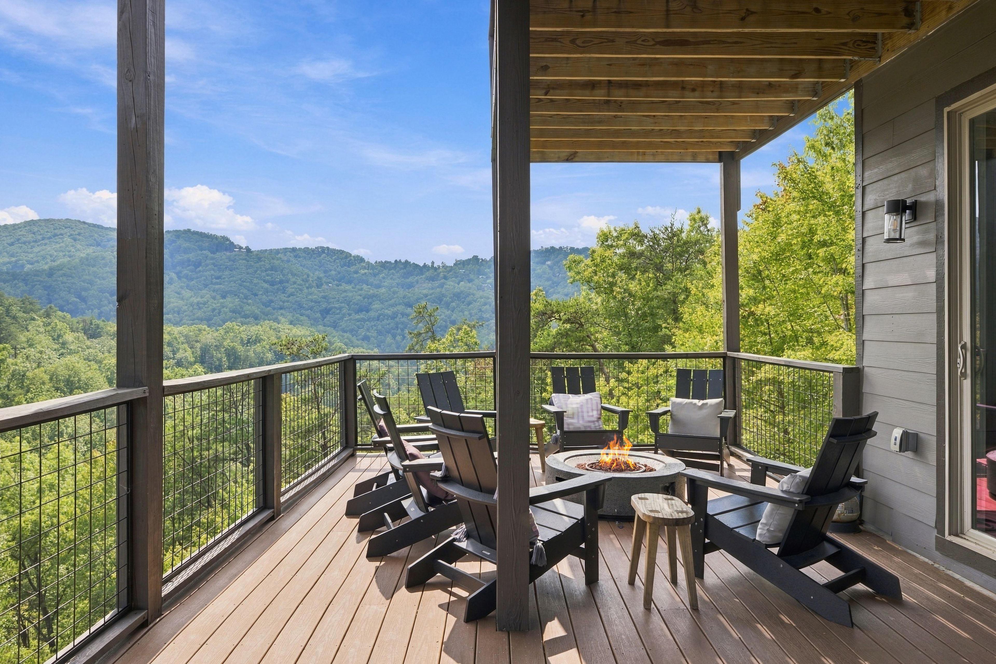 A wooden deck with outdoor furniture and a fire pit, overlooking a forested mountain range.