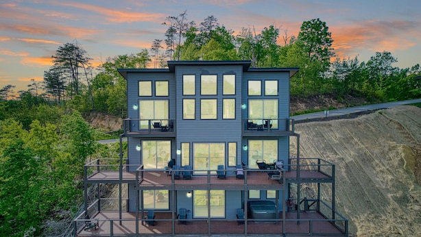 A multi-story building with large windows and outdoor seating areas, surrounded by trees and a rocky landscape. - Panoramic Paradise 4 Bedroom Cabin by RedAwning (Sevierville)