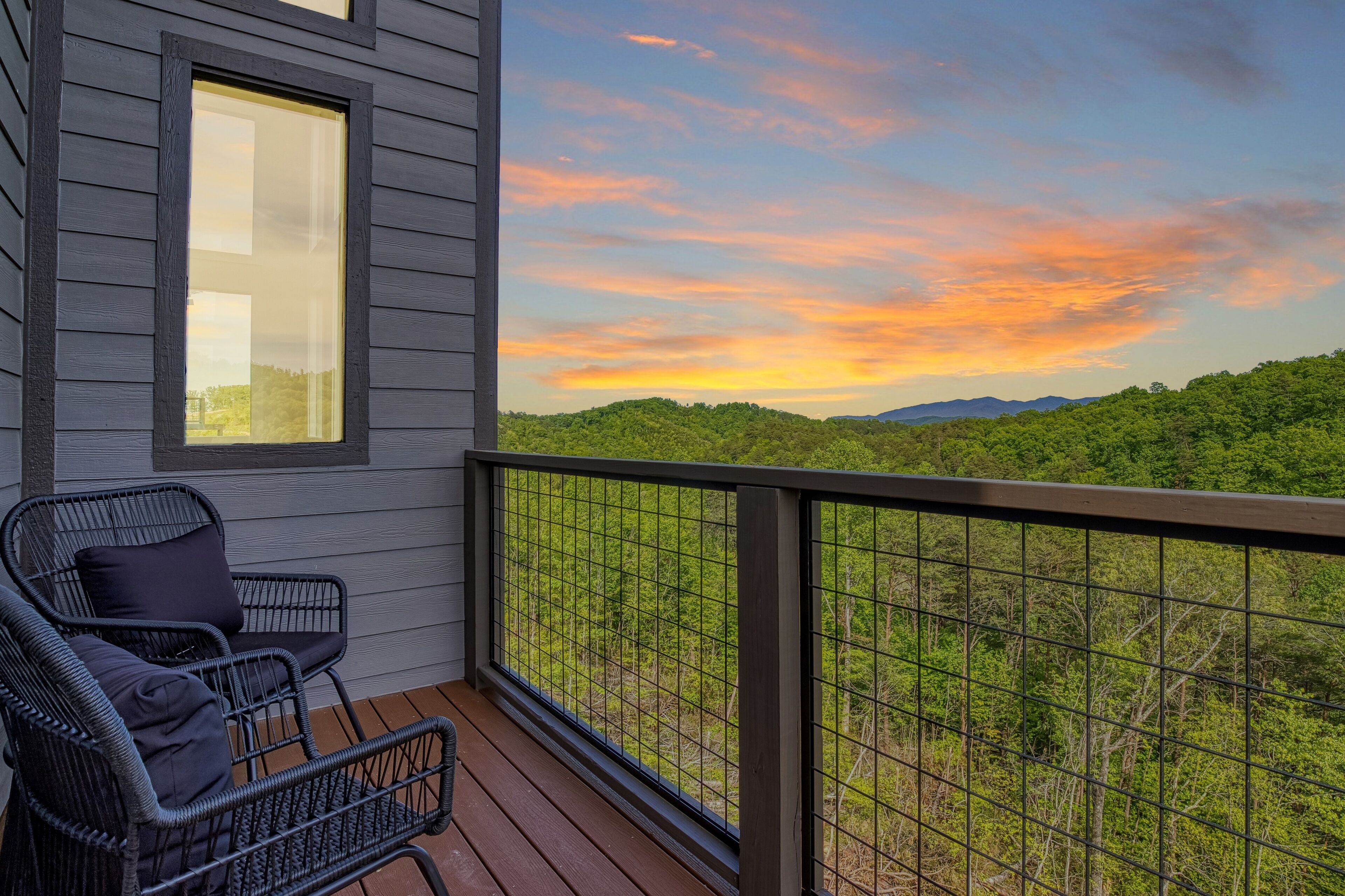 Een balkon met twee rieten stoelen en uitzicht op een bebost landschap in de zonsondergang.