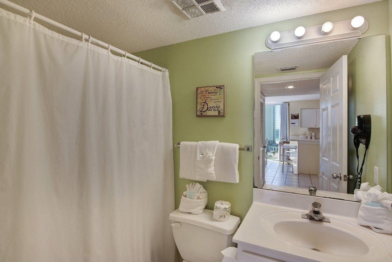 Un baño con cortina de ducha, inodoro con toallero, lavabo con espejo y un cartel en la pared.