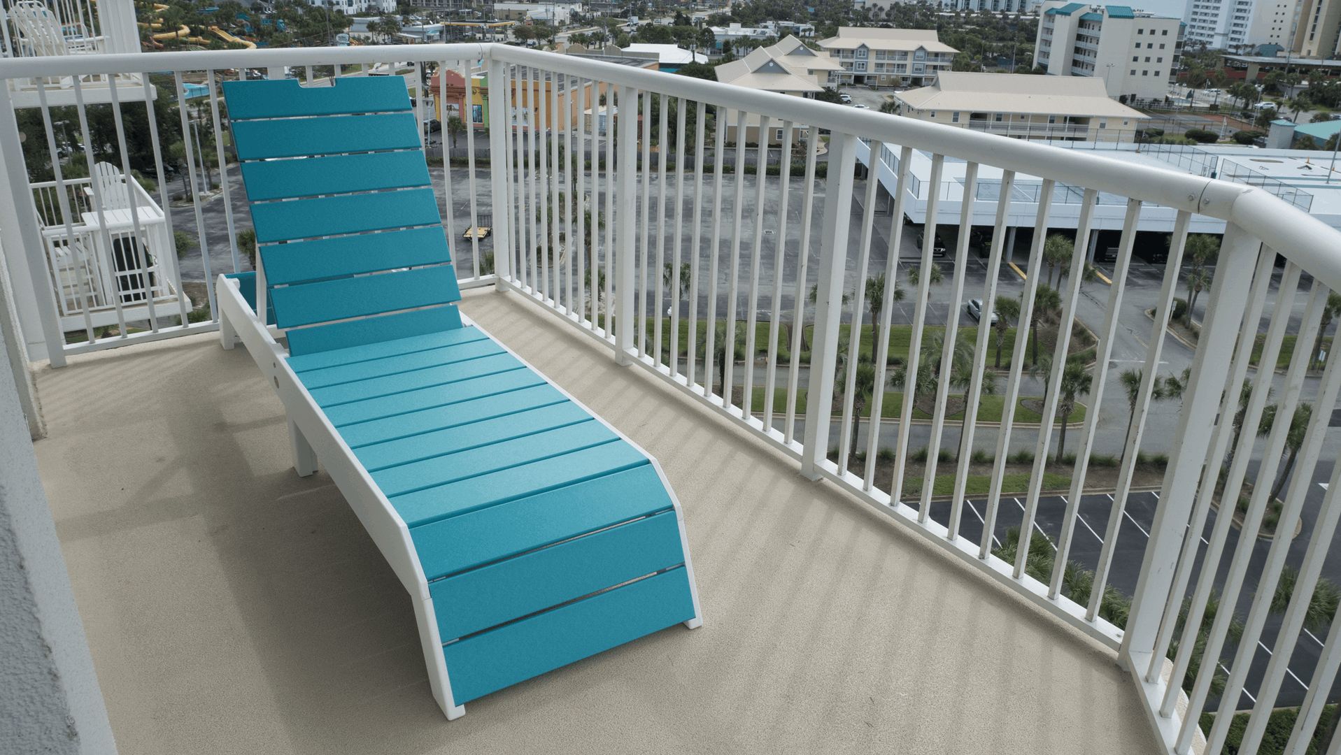 A balcony with a blue lounge chair, white railing, and a view of a cityscape with buildings and roads.