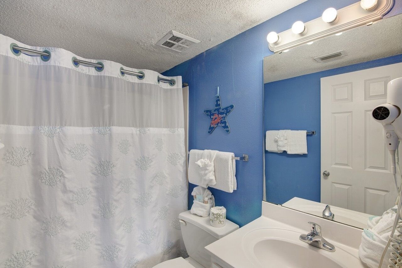 A bathroom with a white shower curtain, a blue wall, a white toilet, a white sink with a mirror, and a hairdryer.
