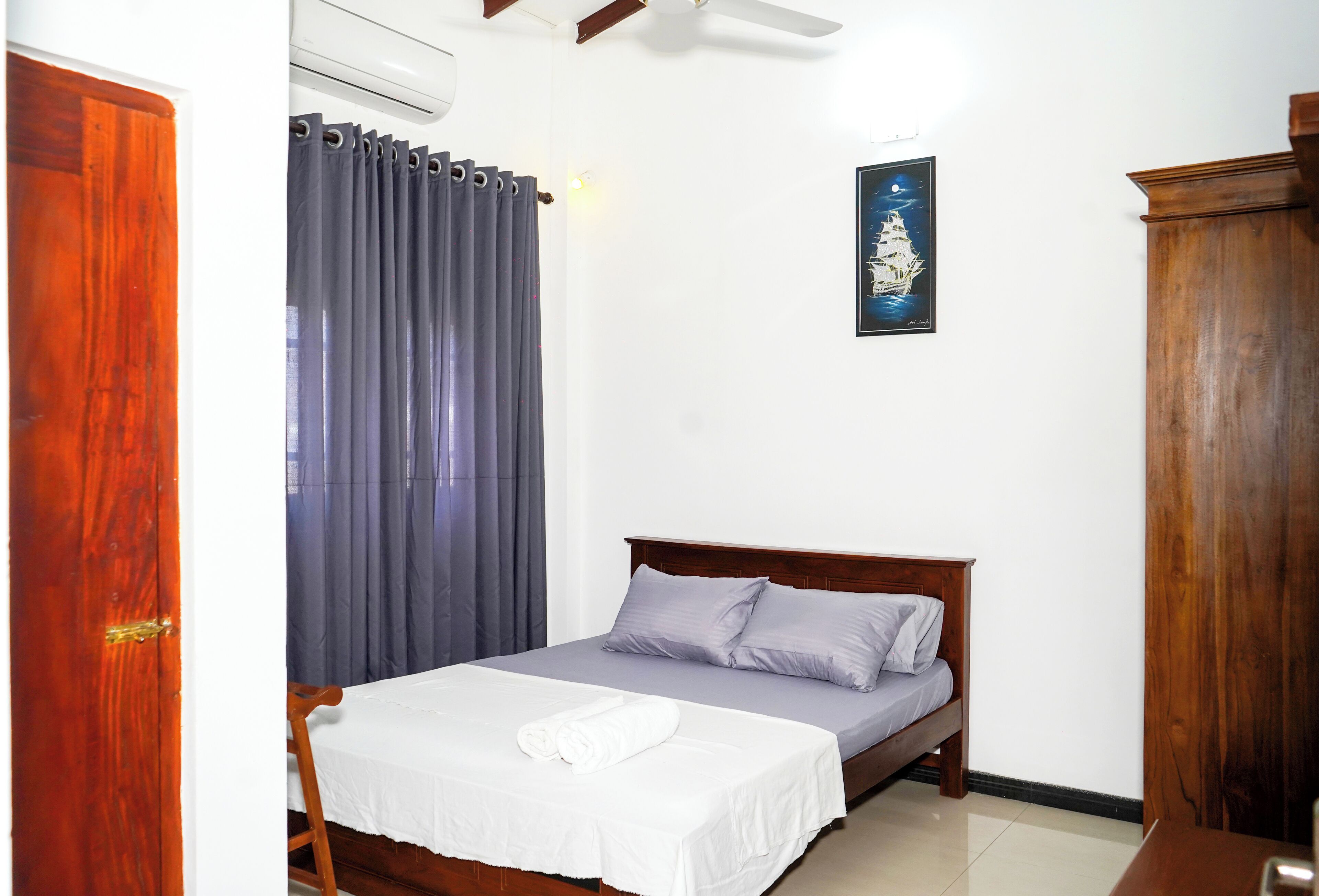A neatly made bed with a white bedspread and two pillows, a wooden headboard, a gray curtain, and a framed picture on the wall.